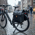 Fahrrad mit Basil Urban Dry wasserdichter Doppelpacktasche matt schwarz im Regen auf nassem Stadt-Kopfsteinpflaster.