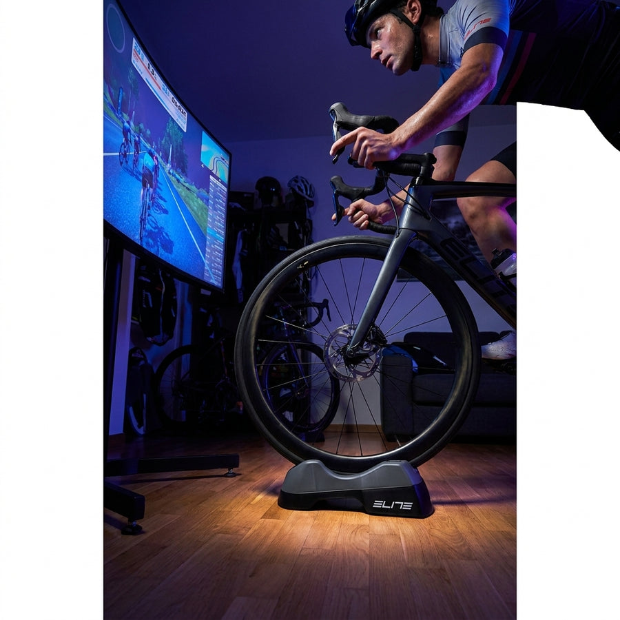 ELITE Sterzo Smart Travelblock Vorderradstütze mit Lenkfunktion für Radfahrer beim Indoor-Training vor großem Bildschirm.