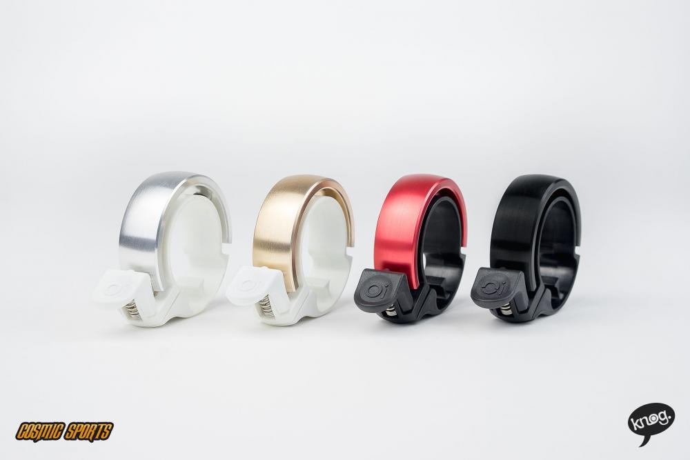 Knog Oi Classic Fahrradklingeln in Schwarz und Rot, auch Silber und Gold, auf weißem Hintergrund.